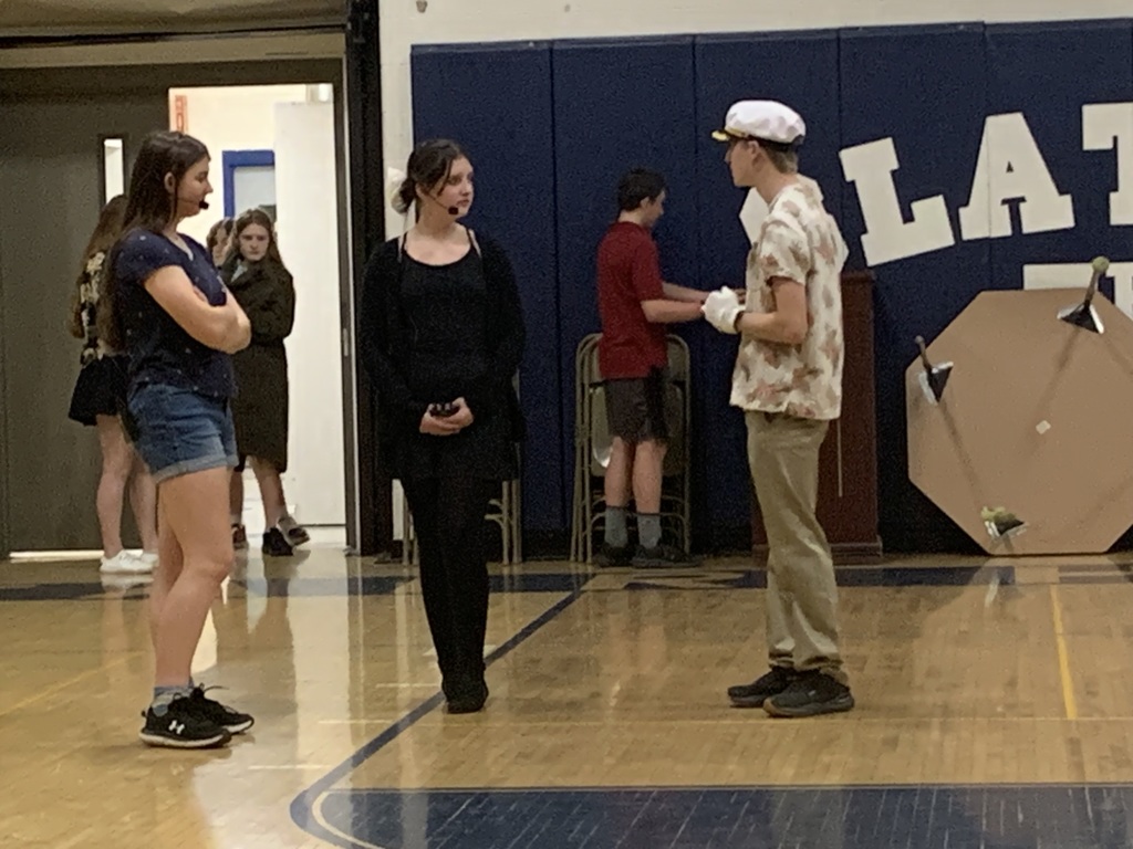 Three students rehearse a scene in a school gym. One student wears a captain’s hat and gloves while speaking to two students standing nearby. Other students and props are visible in the background.
