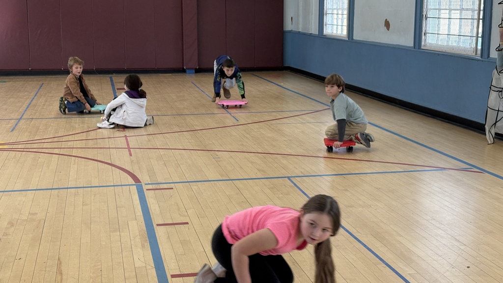 Four children ride small scooter boards around a gym floor. One child in the foreground is on a pink scooter board, while the others use blue and red boards. They appear to be playing a game and are spread out across the gym.