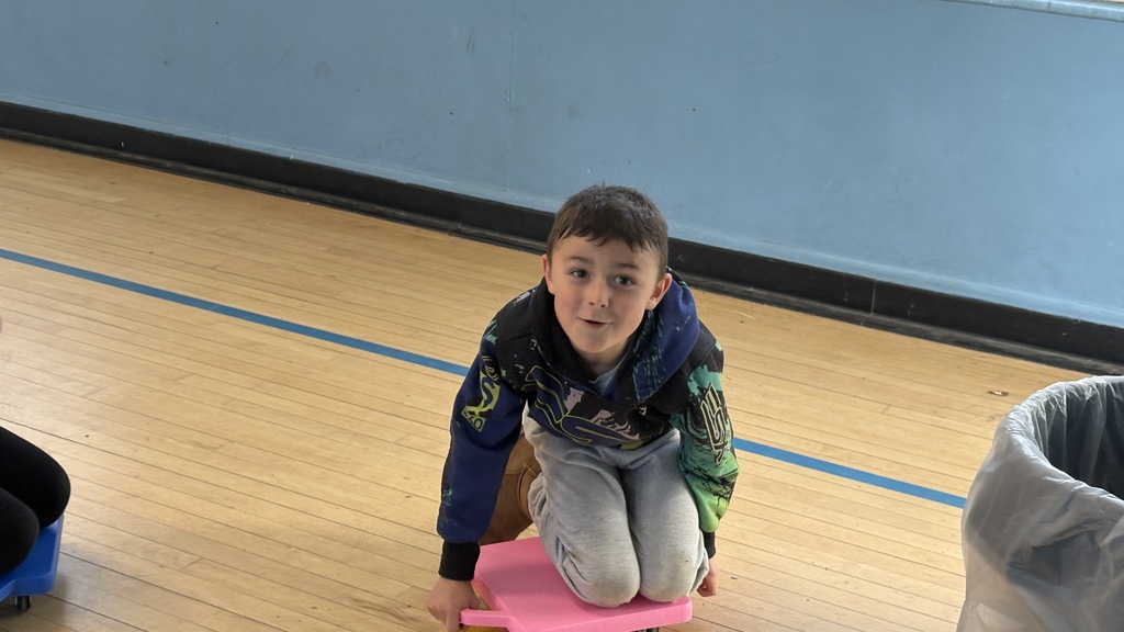A young child kneels on a small pink scooter board inside a gymnasium, looking toward the camera with a playful expression. The gym's wooden floor and light blue wall are visible in the background.