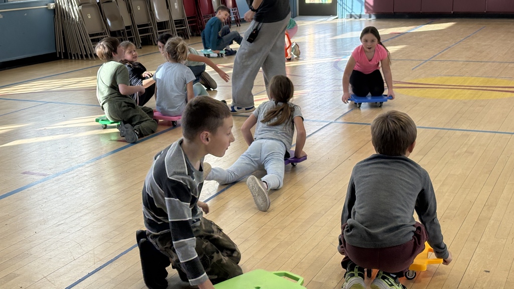 Several children ride colorful scooter boards across a gymnasium. Some are sitting or kneeling on the boards while others push themselves forward. A teacher or adult stands in the background supervising.