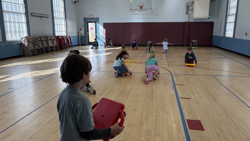 Children ride scooter boards around the gym while one child in the foreground holds a red scooter board and watches. Others are kneeling or sitting on boards as they move across the gym floor.