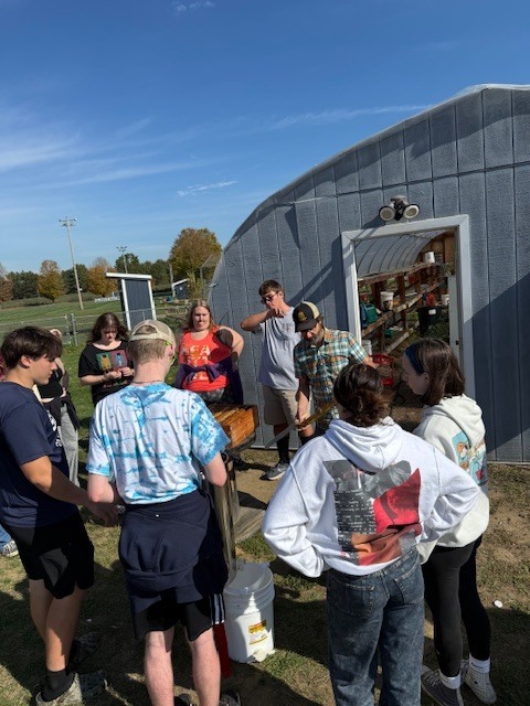 A group of students gathers around a teacher or instructor outside near the beekeeping equipment. A hive box and supplies are on the ground while students watch and listen.