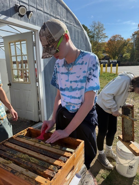 A student wearing a tie-dye shirt and hat holds a honey frame above a hive box while using a brush to remove beeswax. Another student stands nearby holding equipment.