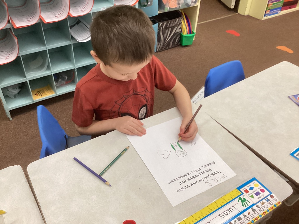 A child wearing a red shirt sits at a desk drawing on a Veterans Day thank-you sheet. Crayons and pencils are spread out on the desk around him.