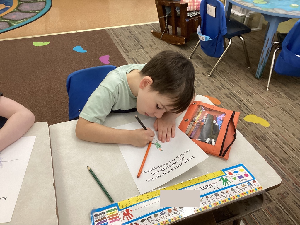 A student leans over a thank-you sheet, carefully coloring a drawing. A pencil pouch and classroom furniture are visible nearby.