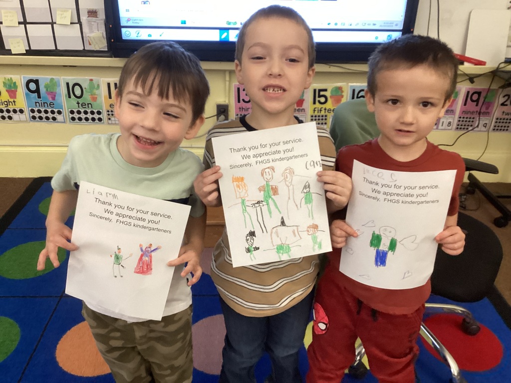 Three kindergarten students stand on a colorful classroom rug, smiling and holding up their completed Veterans Day thank-you drawings.