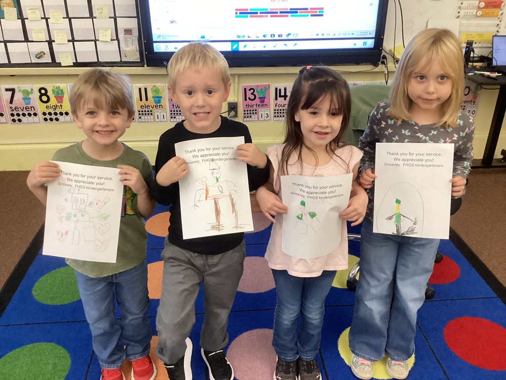 Four kindergarten students stand in a row on a classroom rug, proudly holding up the Veterans Day thank-you drawings they created.