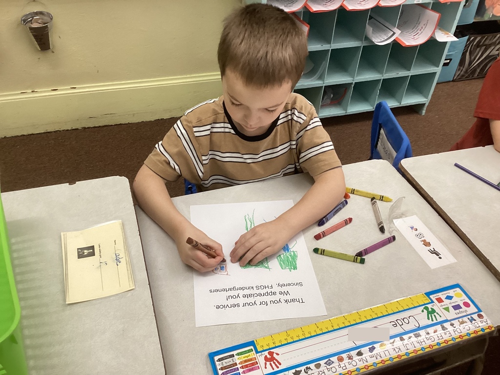 A young student sits at a classroom desk coloring a Veterans Day thank-you sheet with crayons. The desk has a nameplate and scattered crayons, and cubbies are visible in the background.