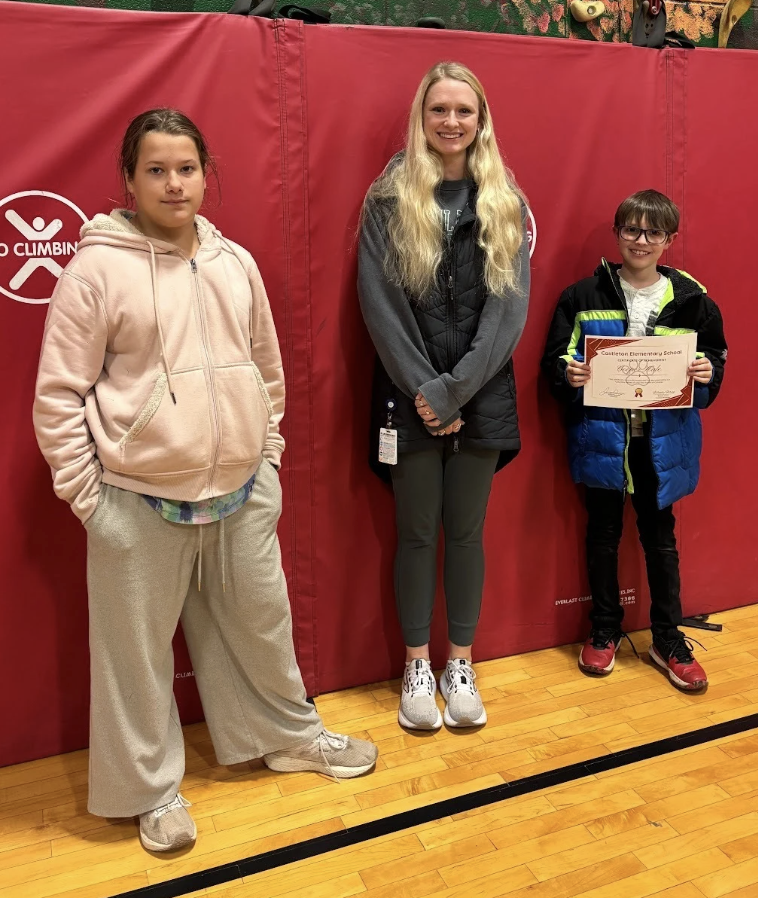 Two students standing with an adult in front of a red gym mat; one student holds a certificate.