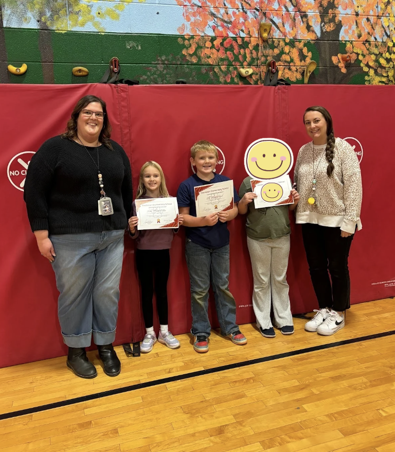 Two students stand between two adults in front of a red gym mat; both students hold certificates.