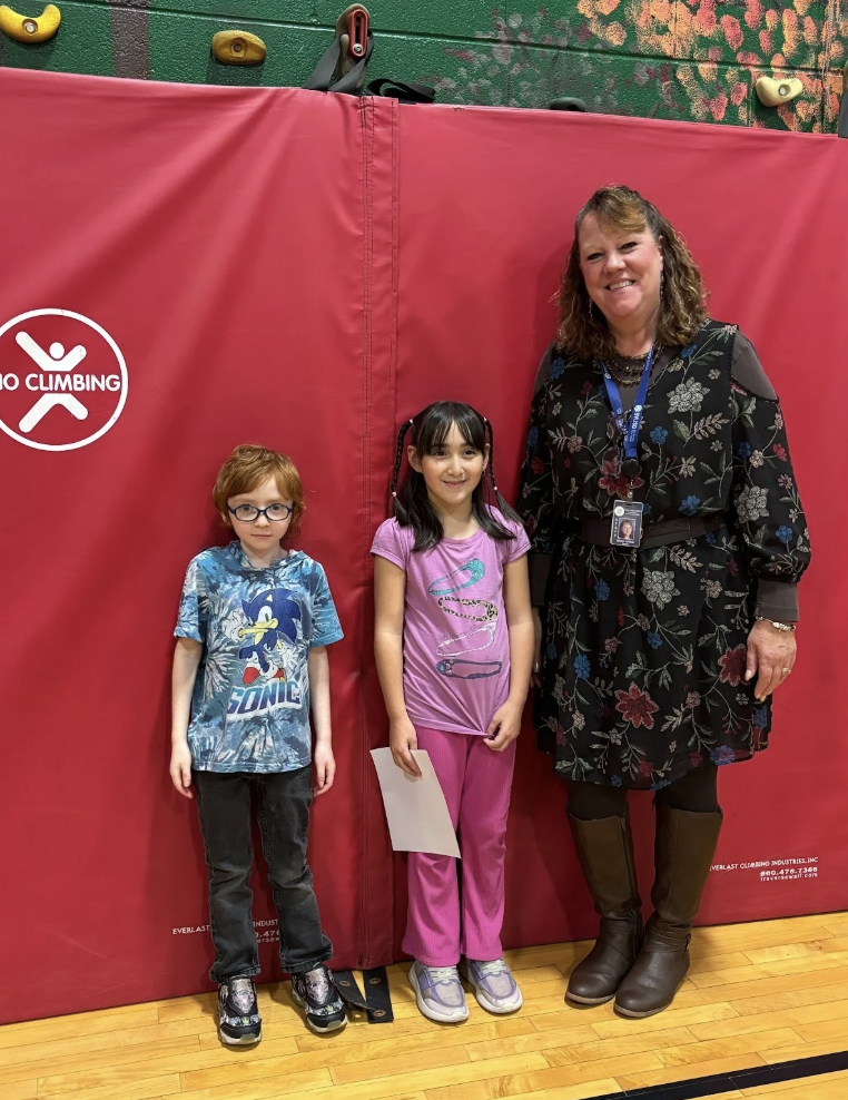 Two students stand with a teacher in front of a mural wall; no certificates visible.