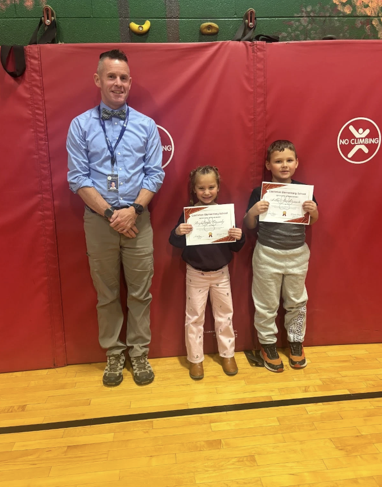 Two students holding certificates stand with an adult in front of a red gym mat.