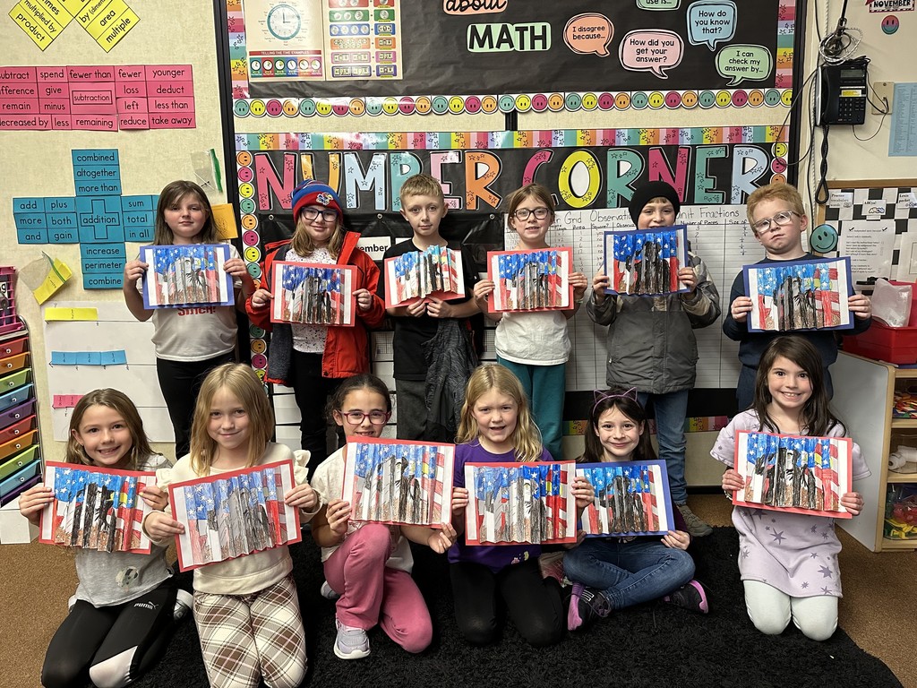 Elementary students stand in two rows in a classroom, each holding artwork of an American flag with an eagle. A colorful classroom math display is behind them.