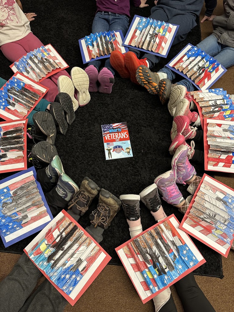 Overhead view of students lying on the floor in a circle with their American flag eagle artwork over their legs; a book titled What Is Veterans Day? is placed in the center of the circle.