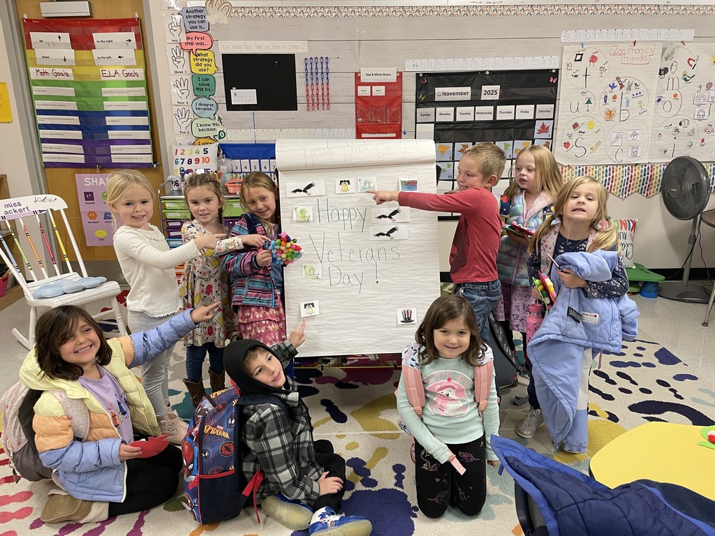 Alt text: A group of young students in a colorful classroom smile and pose around a large chart paper that says “Happy Veterans Day!” The chart includes small pictures of flags, birds, and handprints. The children are wearing jackets and backpacks, and some are pointing to the poster. The classroom is decorated with learning charts, a rocking chair, and bright rugs and materials