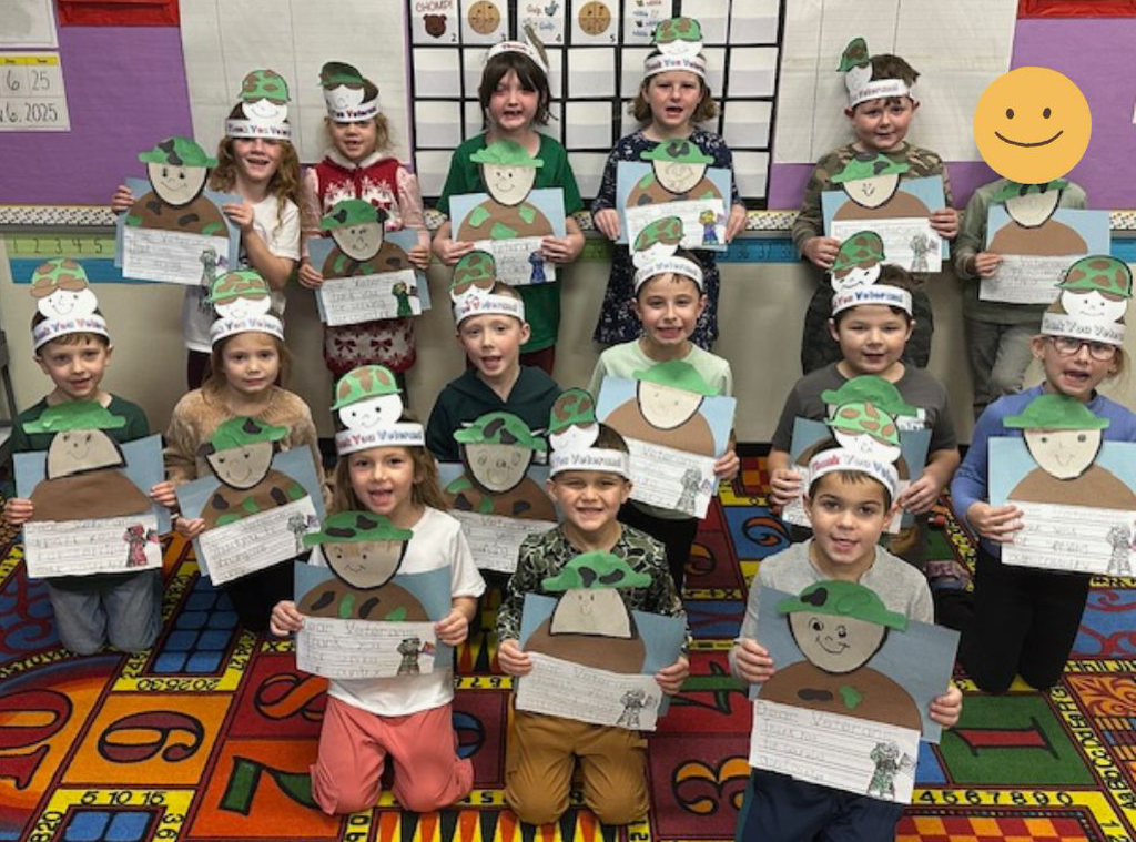 A group of first-grade students from Mrs. Roberts’ class at Castleton Elementary School stand in rows on a colorful classroom rug, proudly holding their Veterans Day art projects. Each project features a paper soldier with a handwritten thank-you note. The students wear handmade hats that say “Thank You Veteran” and smile for the photo.