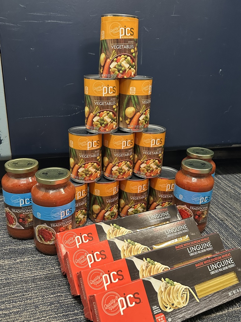A display of food drive donations, including stacked cans of mixed vegetables, jars of pasta sauce, and boxes of linguine, arranged neatly on a carpeted floor.