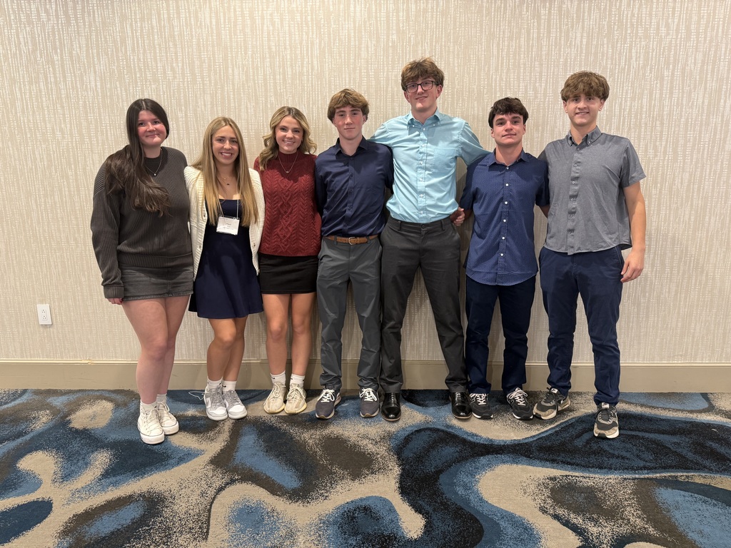 A group of seven students stand side by side in a hotel conference room, smiling for a photo. They are dressed in semi-formal attire, with a mix of collared shirts, sweaters, and dresses, standing against a beige textured wall with a blue and gray patterned carpet.
