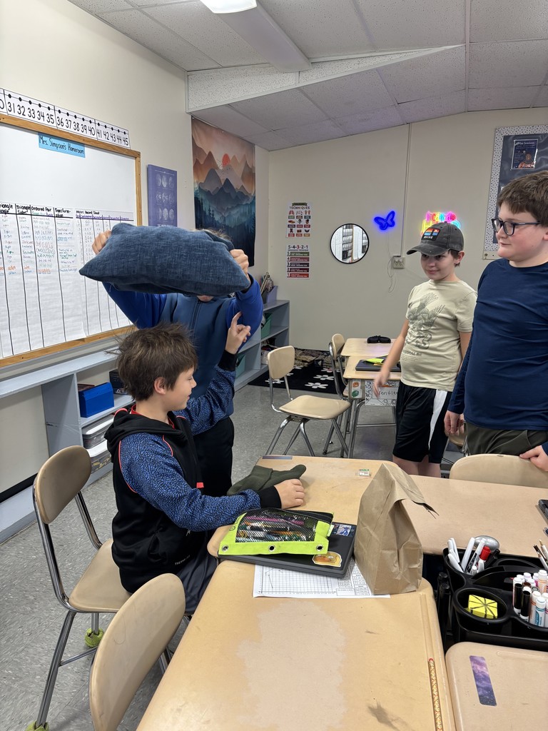 Several students gather around a desk while one student sits, and another holds a pillow above his head during a static electricity activity.
