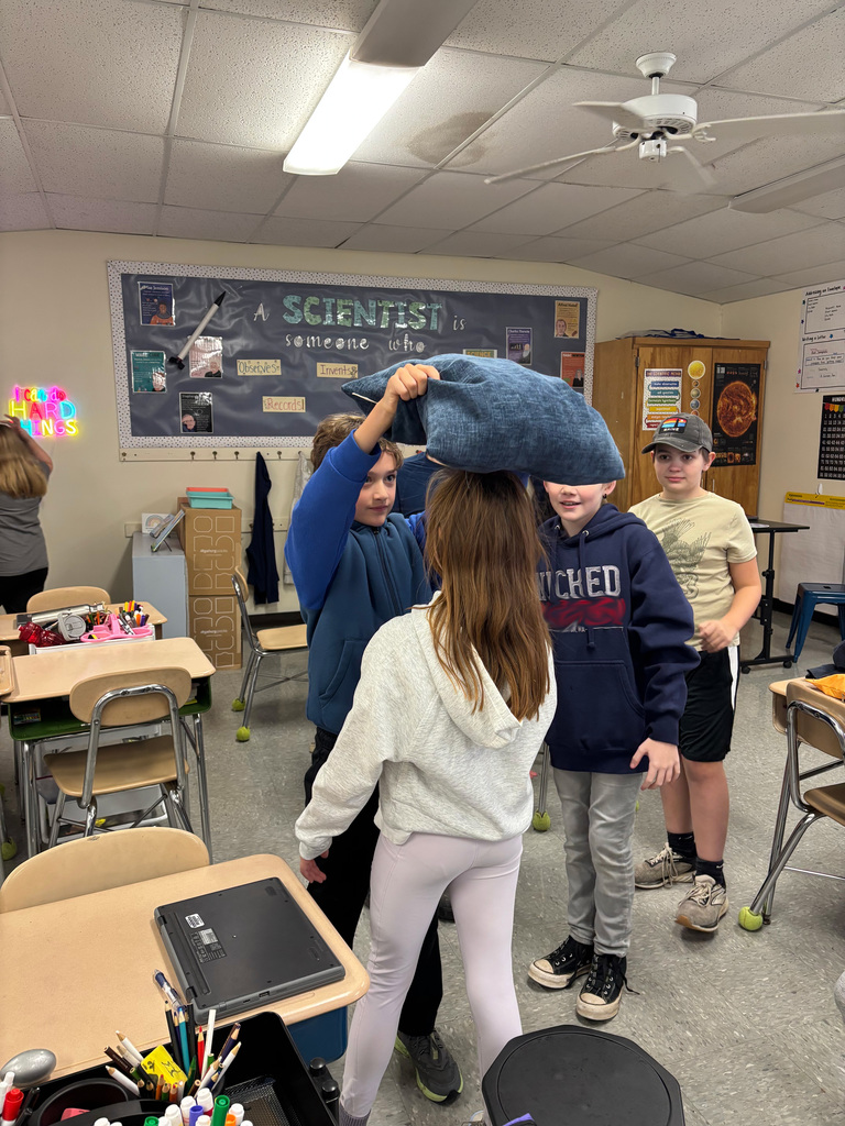Four students in a classroom conduct a static electricity experiment, holding a pillow above a girl’s head to make her hair stand up.