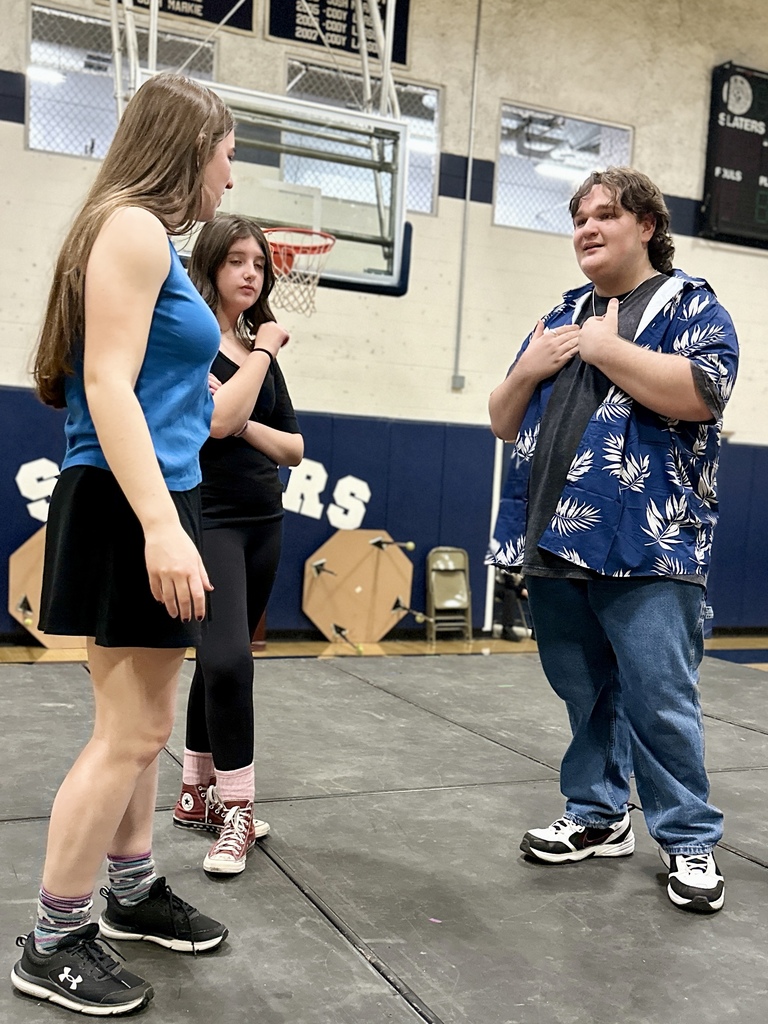 Three students rehearse a scene on stage; one student gestures with both hands while speaking to two others listening.