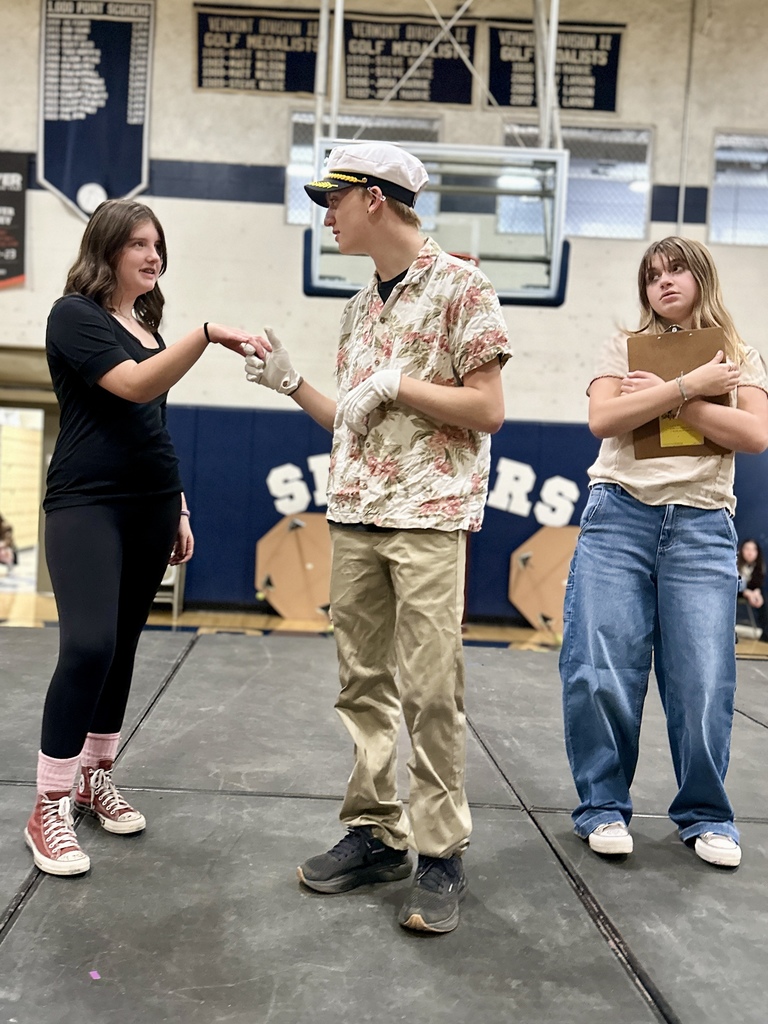 Three students rehearse on stage; one shakes hands with a student dressed as a ship captain while another stands nearby holding a clipboard.