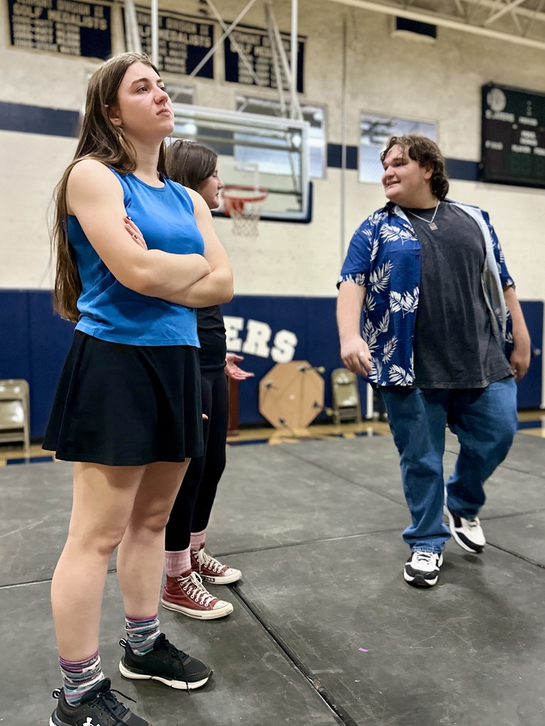 Two students stand in the foreground during a rehearsal, one with arms crossed and the other speaking; a third student in a Hawaiian shirt stands behind them.