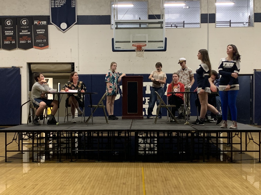 Students rehearse Murder Ahoy on a raised stage in the gym, seated and standing around tables and a podium while practicing a scene.