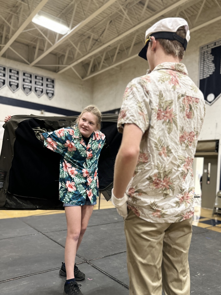 A student opens a long coat to reveal a colorful Hawaiian shirt while rehearsing with another student dressed as a ship captain.