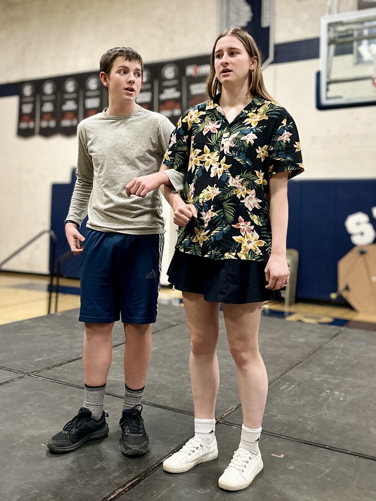 Two students stand on stage rehearsing a scene; one wears a gray shirt and athletic shorts, and the other wears a floral Hawaiian shirt.