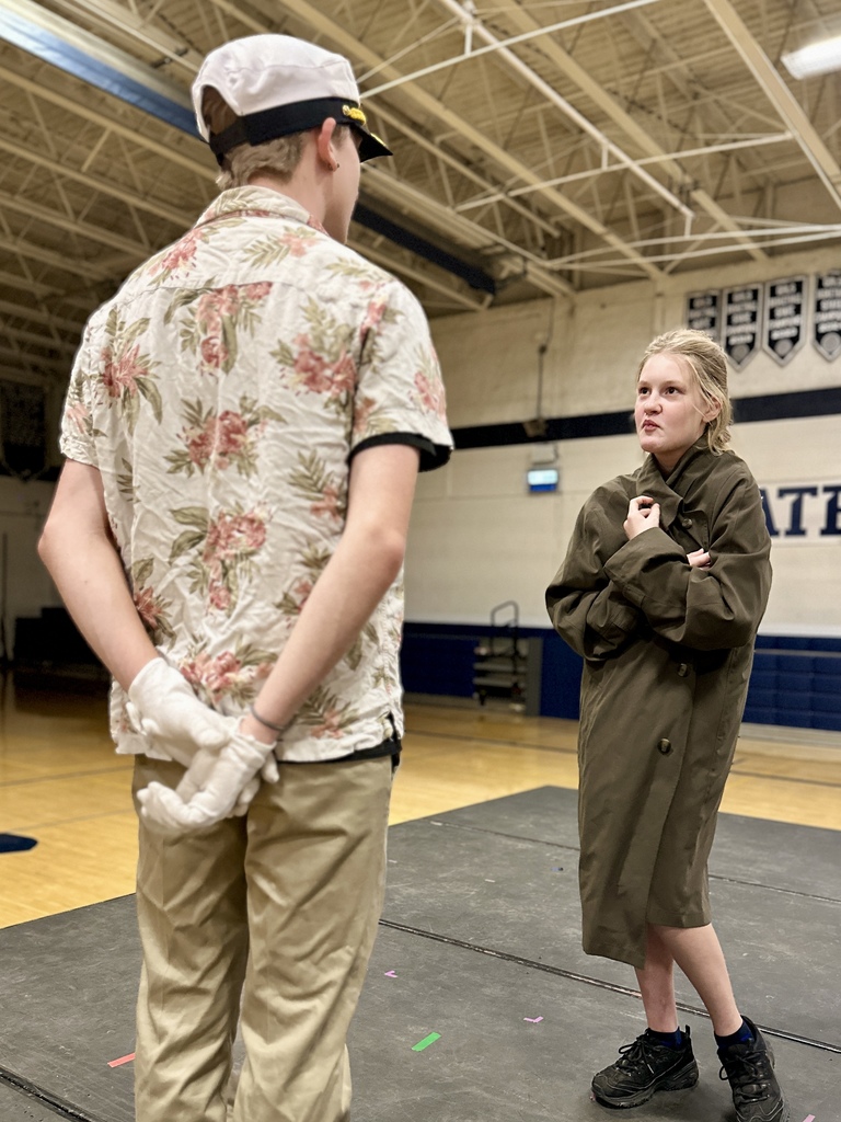 A student dressed as a ship captain faces another student in an oversized coat, acting out a dramatic confrontation on stage.
