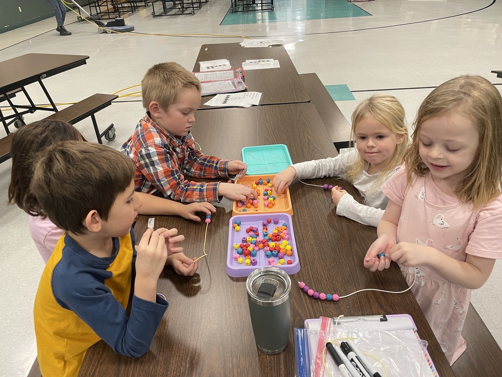 A high-angle close-up of four children gathered around a brown table, using plastic tongs (or scoops) to transfer small, colorful pom-poms between an orange tray and colored plastic cups. They are leaning in intently, focusing on the fine motor skill activity.