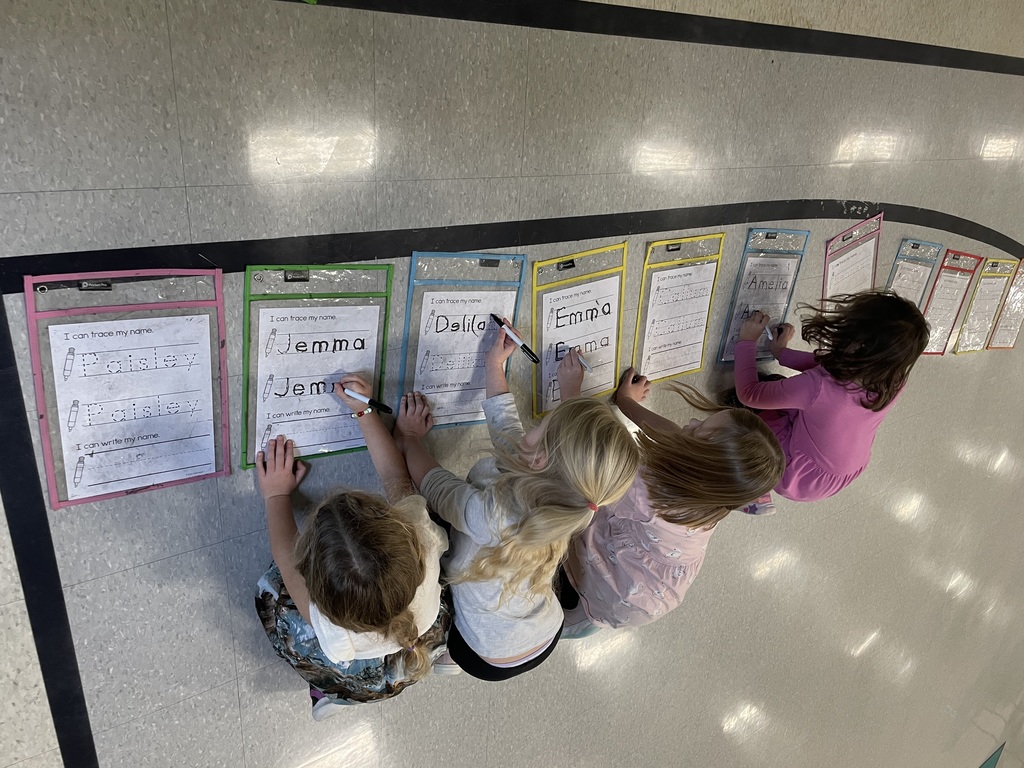 An aerial view of four young girls kneeling along a black line on the floor, tracing their names onto personalized worksheets in colorful laminated sleeves