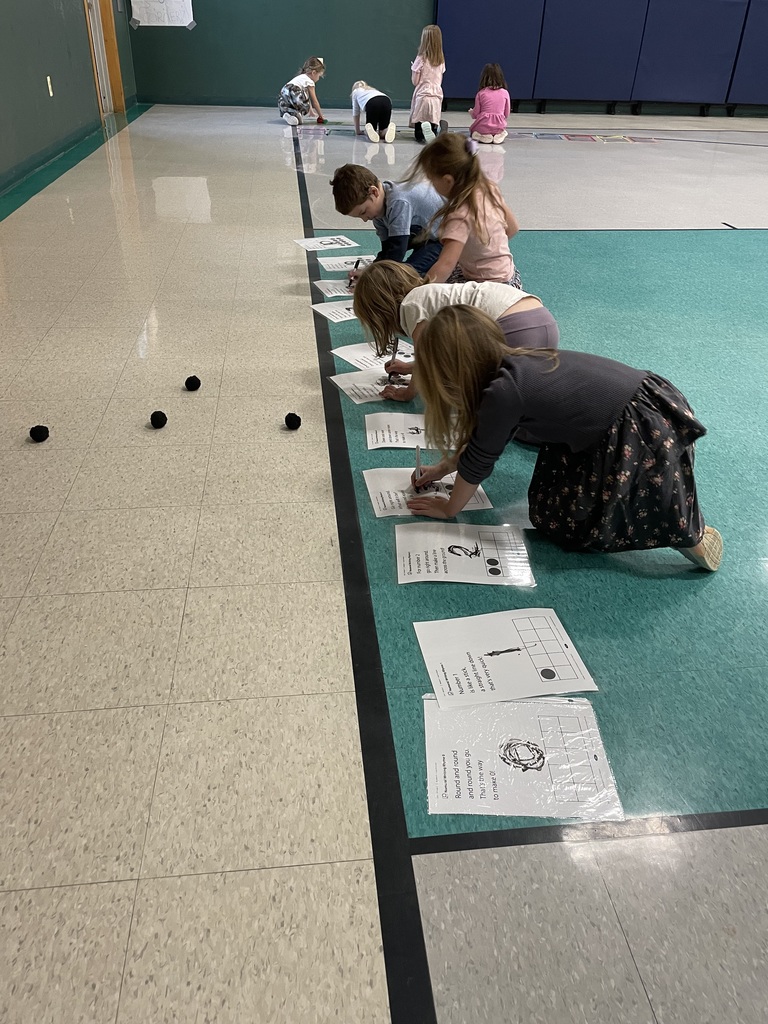 A wide-angle shot showing a group of children working on the floor of a large room (like a gym). In the foreground, four children are kneeling and writing on worksheets laid out in a row. A few black balls are scattered nearby, and more children are visible working in the distance.
