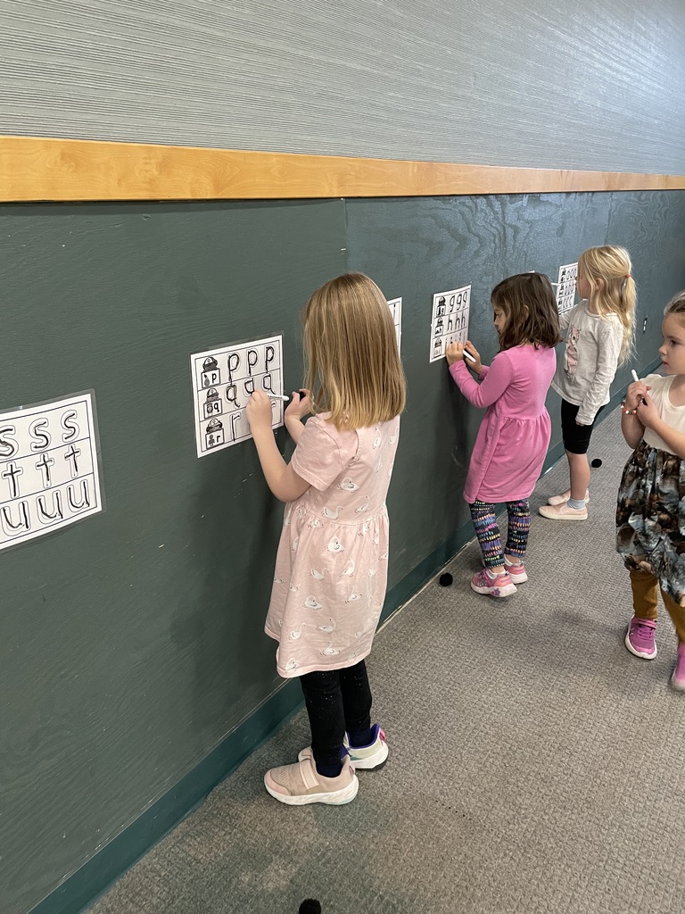 Four young girls are standing side-by-side along a dark green wooden wall, writing with markers on laminated worksheets taped to the wall. The worksheets show large letters (S, T, U) and alphabet tracing activities, indicating a literacy or handwriting exercise.