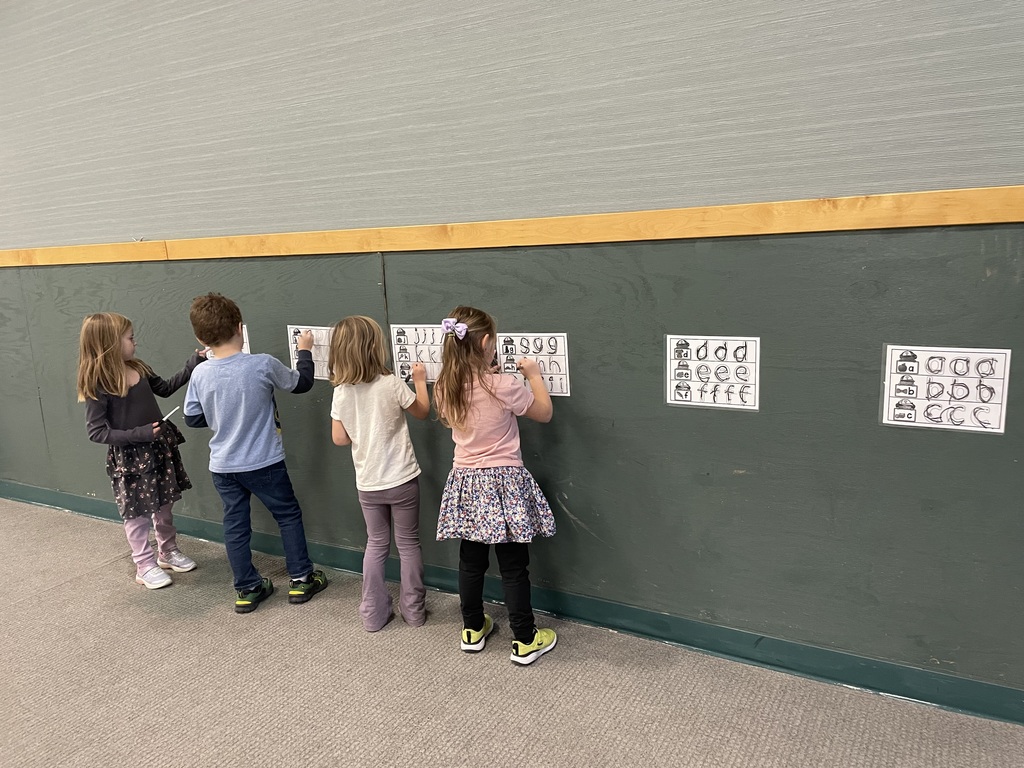 Four young girls are standing side-by-side along a dark green wooden wall, writing with markers on laminated worksheets taped to the wall. The worksheets show large letters (S, T, U) and alphabet tracing activities, indicating a literacy or handwriting exercise.