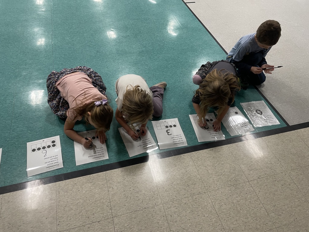 An aerial view of four young children kneeling on the floor, which has a mix of green and tiled sections. They are all bent over, writing with markers on individual worksheets laminated in plastic, which feature number lines or ten frames for counting practice.