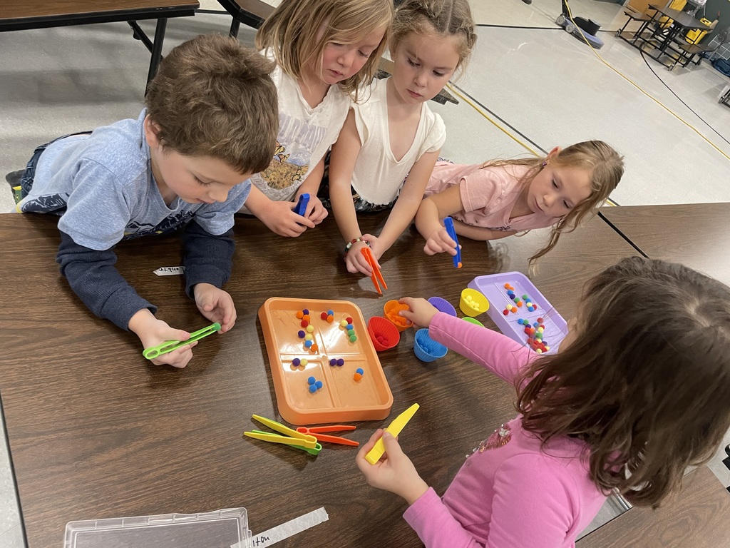 A high-angle close-up of four children gathered around a brown table, using plastic tongs (or scoops) to transfer small, colorful pom-poms between an orange tray and colored plastic cups. They are leaning in intently, focusing on the fine motor skill activity.