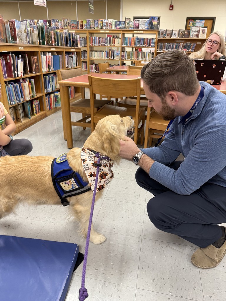 An adult staff member kneels on the floor of the school library, smiling as he pets a golden therapy dog wearing a blue vest and fall-themed bandana. Bookshelves, tables, and a staff member working on a laptop are visible in the background.