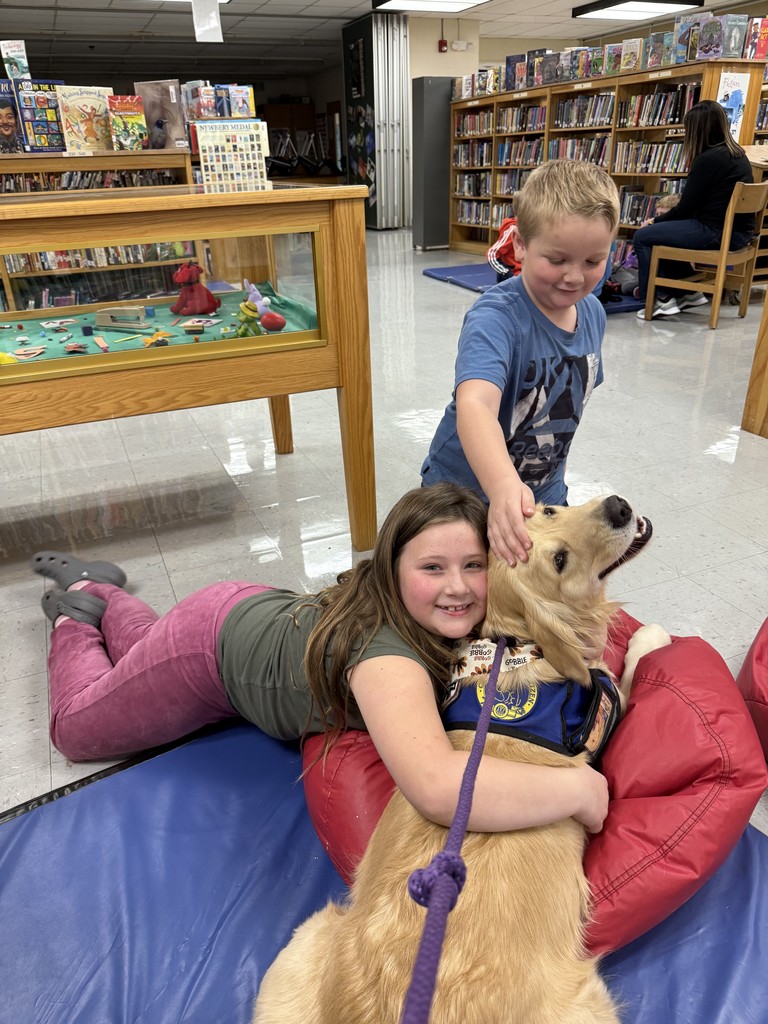 Two students happily cuddle and pet a golden therapy dog on a blue mat and red beanbag chairs in the school library. One child lies next to the dog while another reaches over to pet its head. Bookshelves and a display case with toys are visible behind them.