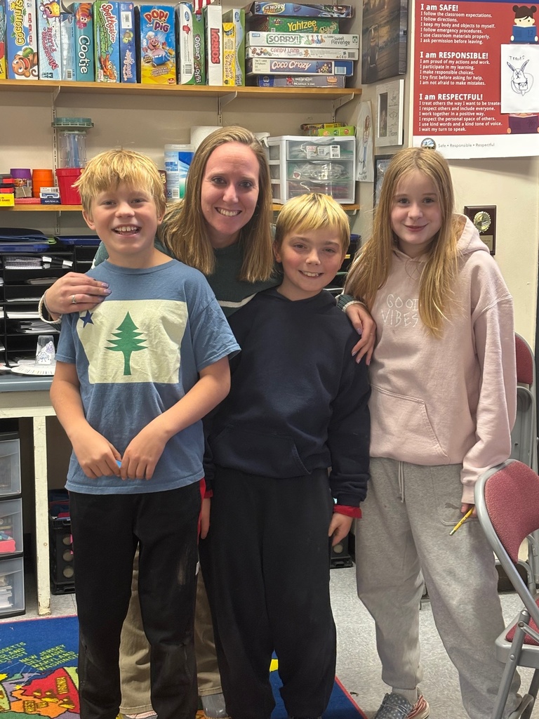 A teacher poses with three smiling students in a classroom. They stand in front of shelves filled with board games, art supplies, and bins. A classroom expectations poster is visible on the wall behind them.