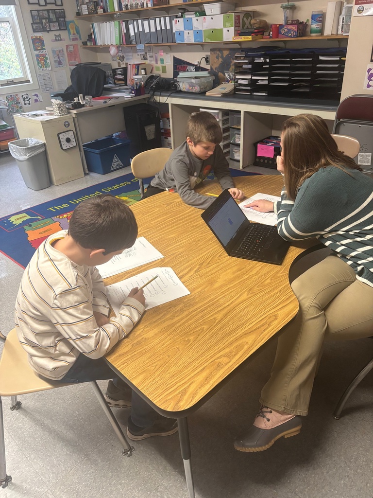 A teacher sits at a kidney-shaped table with two young students, each working on worksheets. One student writes while the other follows along on a chart. The teacher has a laptop open and points to the screen. The classroom background includes shelves, binders, posters, and student artwork.