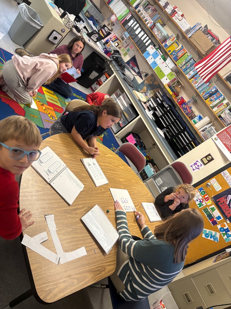 A teacher works with a small group of students at a round table covered with math materials, including number models and cut paper strips. Other students sit or kneel on the classroom carpet nearby, also engaged in work. Shelves, bins, and classroom supplies line the walls.