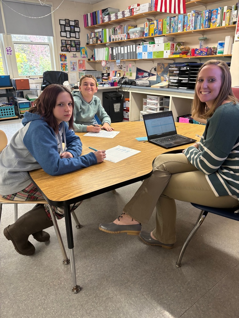 A teacher sits at a small table with two older students who are completing worksheets. All three smile at the camera. The classroom background includes shelves filled with binders, books, and games, along with an American flag near the window.