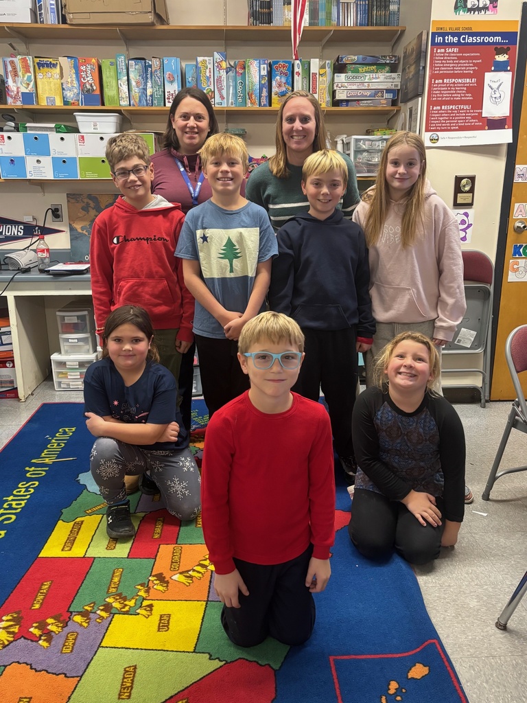 A group photo of two adults and eight students standing and kneeling together on a colorful United States map classroom rug. Everyone is smiling. Behind them are shelves with board games, classroom supplies, and a large expectations poster.