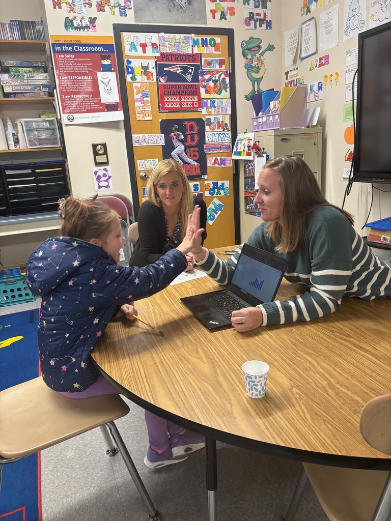 A teacher sits at a table with a laptop displaying a bar graph while a young student in a winter jacket reaches out for a high-five. Another adult sits beside the student, smiling. Behind them is a colorful classroom wall with posters, names, and sports-themed decorations.