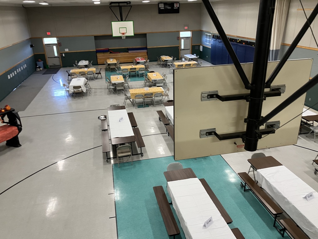 A high-angle view of a gymnasium floor set up for a meal. The room contains rows of tables, some covered with white tablecloths and others with yellow patterned cloths. Long picnic-style tables with benches are visible in the foreground, with time cards (e.g., "11:45") placed on them. The floor is marked with sports lines and a teal blue border.