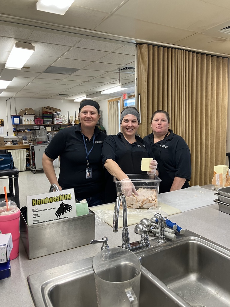 Three cafeteria workers (all women wearing matching black shirts and hair coverings) are standing behind a stainless steel counter in a kitchen or food preparation area. The woman in the center is holding a block of cheese over a clear bin of prepped food. In the foreground, there is a stainless steel sink and a sign with instructions for Handwashing.