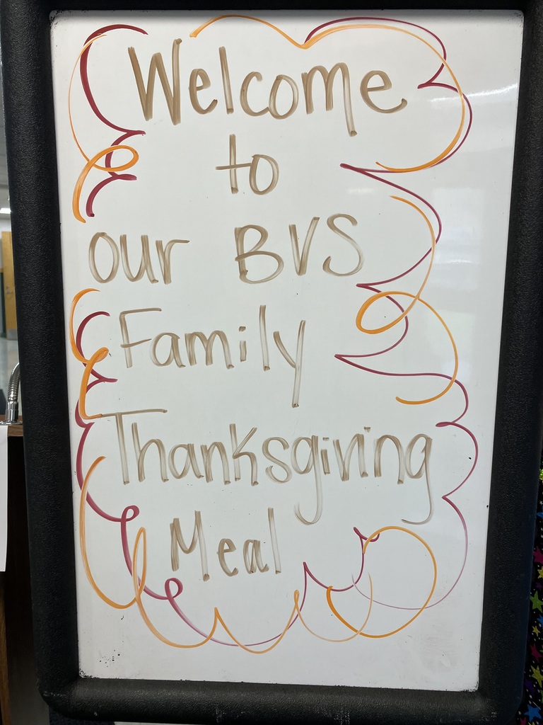 A close-up photograph of a white marker board in a black frame with a hand-drawn message in brown, orange, and red marker that reads: "Welcome to our BVS Family Thanksgiving Meal". The text is surrounded by decorative squiggly lines.