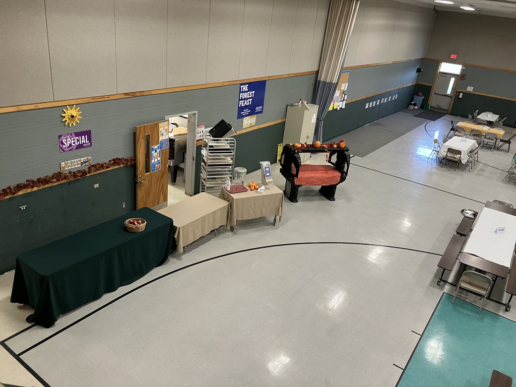 A high-angle, wide shot of the gymnasium set up for the meal, showing the serving and entrance area. Several tables draped in tan and dark green cloths are set up near an open doorway to the kitchen. Fall decorations, including pumpkins and a sign reading "THE FOREST FEAST," are visible along the wall.